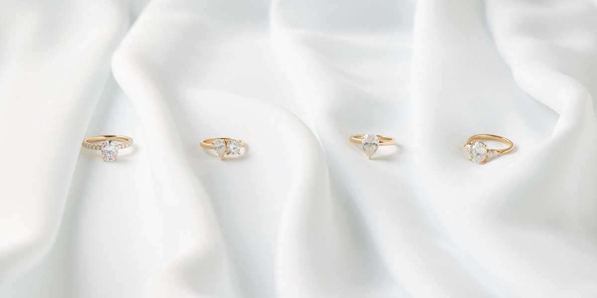 Four gold engagement rings on white silk: an oval with a pavé band, a pear and round toi et moi, a pear solitaire and an oval three stone